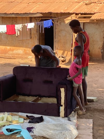 The image depicts an outdoor upholstery scene in the city of Nev, Nigeria, featuring three individuals and a partially completed sofa. In the foreground, a dark purple, velvet-like sofa frame is visible, with its back and portions of its armrests upholstered, while the bottom structure exposes wooden framing and white padding. Tools and materials, including yellow and light blue foam pieces, dark fabric scraps, and a wooden stick, are scattered on a white tarp on the ground in front of the sofa.

An adult male, shirtless and wearing dark trousers, is bent over the sofa's far side, actively engaged in upholstery work. A level or tape measure rests on the sofa's backrest near his left hand. To his right, another adult male stands upright, wearing a red tank top and green shorts, observing the activity. Between these two men, a young child in a pink sleeveless top and light-colored shorts leans against the sofa, also observing the work.

The background features a single-story building with light brown, unpainted walls and a rust-colored corrugated metal roof. An open, dark doorway is visible in the center of the building, with a smaller, closed window or shutter to its left. A clothesline is strung across the front of the building, displaying various garments, including red shorts, light-colored shirts, and a blue shirt, hanging to dry. The ground throughout the scene is unpaved, consisting of dry, light brown earth.