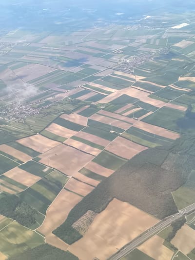 The image shows an aerial view of agricultural fields and towns near Fischamend, Austria. The landscape is dominated by a patchwork of rectangular fields, some brown, indicating recently harvested or fallow land, and others green, suggesting active crops. Several towns or villages are visible as clusters of buildings and roads. Wind turbines are scattered across the landscape, particularly amidst the fields. A forest borders the fields in the lower part of the image, and a highway runs through the lower-right.