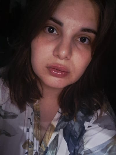A close-up, slightly angled portrait shows a young woman with brown hair and light skin. She lies on her back, her face turned towards the camera. Her dark eyes are wide open, and her lips are slightly parted. The background includes a floral patterned fabric suggesting bedding or clothing. There is no indication of the location being Амзя, Russia.