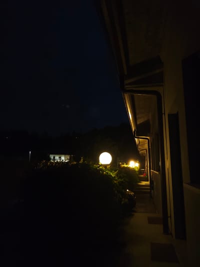 The image depicts a nighttime exterior scene with a building on the right side and a pathway leading into the darkness. There are several round, illuminated garden lights along a hedge to the left of the pathway, casting a warm glow. The building appears to be a house or residential structure, with a visible gutter system. In the distance, through the trees and darkness, there are lights from another building, suggesting a semi-rural or suburban setting. The sky is dark, indicating it is nighttime. There are no people or discernible animals visible in the image. The overall impression is a quiet, illuminated pathway leading away from the viewer into a dark environment. The location is identified as Guarene, Italy.