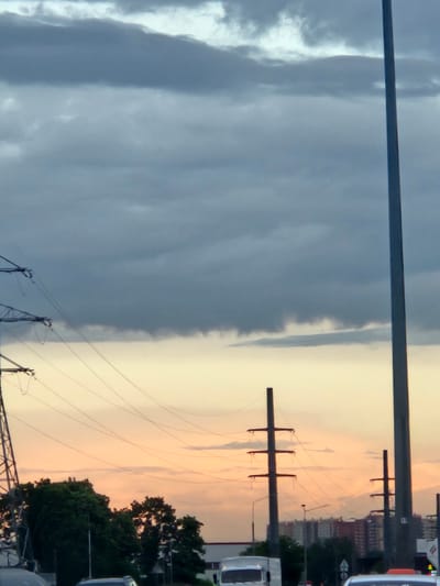 The image shows a scene in Tomilino, Russia, at dusk or dawn. A sharp weather front divides the sky, with dark, stormy clouds on the left and a pale, orange-tinged sky on the right. Power lines and pylons stretch across the sky. Buildings are visible in the background on the right. Vehicles are also present on the ground on the right.