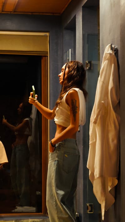 The image depicts a young woman in an indoor setting, identified as being in Mu Cang Chai, Vietnam. The woman, shown in profile, possesses long, dark, wavy hair, with some strands styled into braids or twists. She is wearing a white sleeveless crop top that exposes her midriff and light blue, loose-fitting jeans. A dark, stylized tattoo, resembling a plant or abstract design, is visible on her left upper arm. She holds a smartphone with a prominent green case in her right hand, extended forward towards a large mirror to her left, indicating she is taking a selfie or video. Her left hand is casually placed in her jeans pocket, and a dark bracelet adorns her left wrist.
The immediate background consists of dark, likely grey or blue-grey, textured walls. Architectural features such as recesses or columns are visible on these walls. To the far left, a wooden frame, possibly for a window or door, is visible. The top portion of the scene shows a dark ceiling with wooden beams or paneling elements. On the wall to the right of the woman, a white, textured bath towel hangs from a metal towel rack. The entire scene is dimly lit, suggesting an intimate or low-light environment. The woman's reflection is clearly visible in the mirror, mirroring her posture and action.