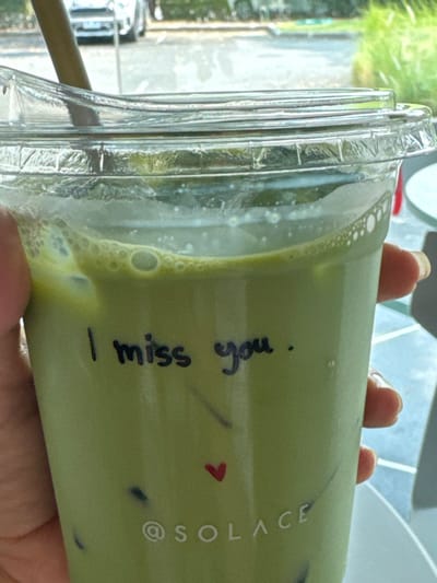 A translucent plastic cup of matcha latte with ice is held in someone's hands. The drink is a pale green color with foam at the top. Written on the side of the cup in black marker is the phrase "I miss you," with a small red heart drawn below. Printed vertically is "@SOLACE." The cup has a ridged lid with a drinking straw visible. A window is visible through the cup, showing greenery and a parked car.