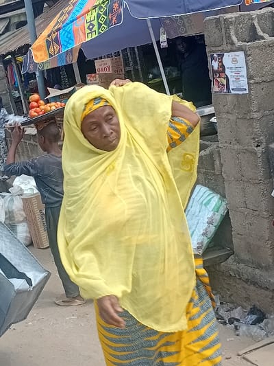 A woman wearing a yellow headscarf stands in a market setting. She has a patterned dress with yellow and grey-blue stripes. A man next to her hands her something while standing beside a display of oranges. Nearby are various goods, including fabrics with colorful patterns and boxes, positioned within what appears to be a bustling commercial area.