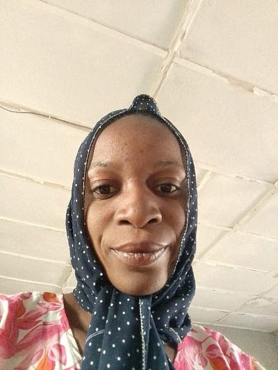 An adult female, likely of African descent, is depicted in a close-up, head-and-shoulders portrait, likely a selfie, in Barnawa, Nigeria. She faces forward, looking directly into the camera with a slight smile. Her skin is dark-toned. She wears a dark blue headscarf with small white polka dots, tied atop her head, framing her face. A pink, white, and orange floral patterned garment is visible at her shoulders and chest. The background is an off-white or light-colored panelled ceiling with visible seams and minor wear, featuring a thin dark wire running horizontally across the upper left quadrant. The lighting suggests an indoor setting.