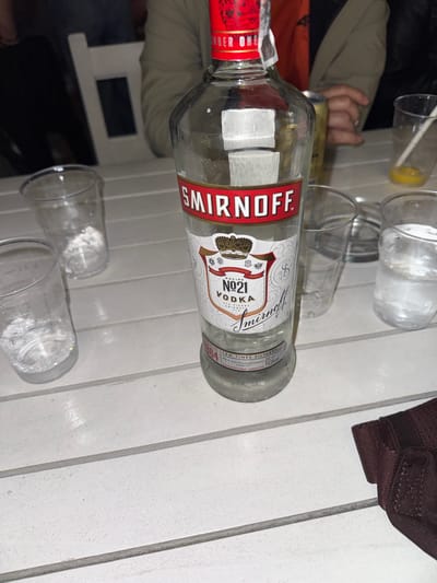 A Smirnoff No. 21 Vodka bottle is the central subject of this image, positioned on a white slatted table outdoors. The bottle is partially empty, with condensation visible on its surface. Surrounding the bottle are several clear plastic cups, some containing ice and liquid, and one with a yellow liquid and a straw. The table is set on an outdoor surface, suggesting a gathering or party. In the background, parts of chairs and people are visible, indicating a social setting. The presence of a person’s hand reaching towards a can in the background hints at ongoing activity. The environment is casually arranged, suggesting a relaxed social occasion. The image appears to be taken in Obrochishte, Bulgaria.