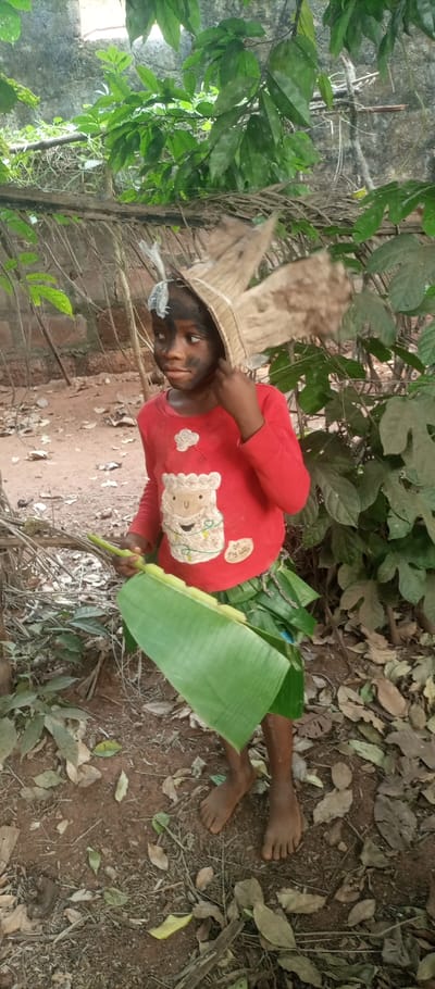 A young person with darkened facial makeup is in a wooded area. They wear a red shirt with a sheep graphic and a leafy skirt. A headdress of straw or dried leaves adorns their head. They hold a large green leaf in their hands, possibly a banana leaf. The ground is dirt-covered and scattered with fallen leaves. Green foliage surrounds the person, suggesting a natural or forest setting. The overall impression is of a costume or role-playing scenario.