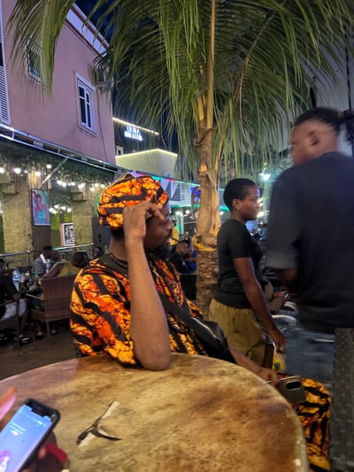 A group of people are gathered at an outdoor seating area, likely a restaurant or bar, at night. The location is identified as Oba, Nigeria. In the foreground, a man with dark skin, wearing an orange and black patterned headwrap and a matching shirt, is seated and looking to his right. He has a beard and is wearing a necklace. A cell phone is visible in his hand. In the mid-ground, a woman with dark skin and short hair, wearing a black t-shirt and khaki pants, is also seated and looking to her left. To her right, another person, whose face is not visible, is wearing a black shirt and appears to be interacting with the woman. In the background, other patrons are seated at tables, and the area is illuminated by string lights. Palm trees are present, suggesting a tropical or subtropical environment. The overall scene depicts a casual, social gathering.