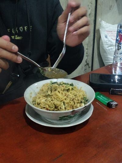 This candid, eye-level shot captures a person eating a bowl of instant noodles in what appears to be a casual indoor setting, likely a small eatery or *warung* in Dramaga, Indonesia. The dominant subject is a person, visible from the mid-torso upwards, wearing a black hoodie with partially visible white-green text that reads "F*CKOFF". Their hands are engaged in the act of eating, using a spoon and a fork to scoop noodles from a white ceramic bowl.
The main dish is a generous serving of instant noodles, seemingly fried or dry-style (*mie goreng*), mixed with bits of green leafy vegetables (possibly bok choy or similar) and scrambled egg. The bowl has a subtle floral design around its rim and rests on a matching white saucer.
On the wooden table, alongside the noodle bowl, are several personal items. To the right, a black smartphone rests on its side, next to a clear plastic water bottle with a blue and red label. Further to the right, a black pack of "MAGNUM" "BINTANG" cigarettes and a bright green lighter are visible. Car keys hang from the left side of the table, near the person's left hand.
The background is simple, with a light-colored, somewhat textured wall on the right side and a large white sack, possibly containing rice or other dry goods, leaning against it. The lighting is soft and ambient, suggesting it might be evening or night. The overall scene conveys a common, everyday meal experience in Indonesia.