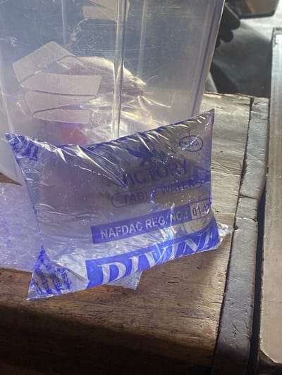 The image shows a plastic sachet of purified water lying on a wooden surface. The sachet is partially transparent and filled with water, with blue text printed on it. Visible text includes "VICTORY TABLE WATER" and "NAFDAC REG. NO.: 01-17...". The sachet appears to be slightly crinkled. The background consists of a rustic wooden table and some other plastic containers. The lighting suggests it might be indoors or in a shaded area. The scene is likely set in Barakin, Nigeria, as indicated by the location context. There are no people or animals visible in the frame, and the focus is on the product. The overall impression is a close-up, still-life shot of a common consumer item.