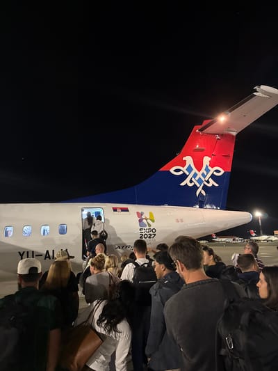 An evening photograph captures a group of passengers boarding an ATR 72 aircraft at Nikola Tesla Airport in Surčin, Serbia. The tail of the plane features a red and blue design with a stylized symbol and the "EXPO 2027 BEOGRAD" logo. Passengers, many carrying luggage, are seen ascending the airstairs into the aircraft. The airport tarmac is visible in the background, with other aircraft parked further away. The scene is illuminated by artificial lighting, casting long shadows and creating a night-time atmosphere.
