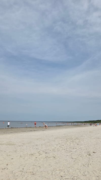 The image depicts a wide, sandy beach in Jūrmala, Latvia, under a cloudy sky. The sea is calm, with shallow water extending a considerable distance from the shore. Numerous people are scattered across the beach and in the water. In the foreground, the sand is textured with footprints and debris. Several individuals are visible closer to the water's edge, including a person in a white shirt and dark pants standing, a child in a blue outfit, and a person in an orange life vest. Further back, more people are wading in the water or relaxing on the sand. The background shows a dense line of trees forming the treeline of the coast. The overall scene suggests a leisurely day at the beach with a moderate number of visitors.
