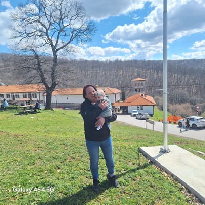 A woman holds a baby in front of the Gigintsi Monastery, Bulgaria. The monastery complex consists of multiple buildings with red tile roofs and a tower. A large bare tree is visible on the left. Behind the complex is a wooded hillside. The sky is blue with scattered clouds. A road with cars and an orange construction barrier runs along the right side. A concrete base for a light post is visible in the foreground, near a grassy area. Other people are at a picnic table.