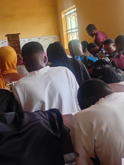 The image depicts an indoor scene, possibly a classroom or meeting hall, with several individuals seated. The room has yellow walls and a window with barred panes. In the foreground, the backs of people wearing predominantly white and dark clothing are visible, suggesting they are facing away from the camera. A lectern is visible on the left side of the image with some writing on it. On the right, a man in a maroon shirt is standing and looking down. The overall atmosphere suggests a gathering or a lecture. This scene is located in Ogbogbo, Nigeria.
