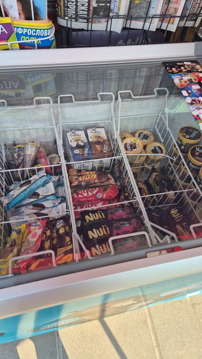 This is a high-angle, medium shot of an open freezer display filled with various ice cream products. The freezer is divided into several compartments, each holding different brands and types of ice cream. In the upper left section, there are empty paper cups. To the right of the cups, there are several packages of "Familia GALAXY" ice cream, with "VANILLA" and "COCOA" flavors visible. Below these, a compartment contains several "Lion" branded chocolate bars and "KitKat" chocolate bars. Further down, there are multiple packages of "Nuii ICE CREAM ADVENTURE" with "DARK CHOCOLATE & NORDIC" flavor. In another section, to the right, there are several round containers of "Nirvana" ice cream, with flavors like "CARAMEL" and "CHOCOLATE & CHEESE CAKE" visible. The freezer is positioned outdoors, as indicated by the concrete ground and the sunlight casting shadows. In the background, above the freezer, there are shelves displaying magazines and newspapers. The overall impression is of a kiosk or small shop selling cold treats, likely in a public area of Ravda, Bulgaria, given the presence of Bulgarian text on some of the packaging and the overall setting.