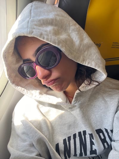 A young person wearing a light gray hooded sweatshirt and oversized purple sunglasses is looking down and to the side. The person's head is partially obscured by the hood. Sunlight is coming in from the left, casting shadows. In the background, a yellow surface with some text and a black object are visible. The scene appears to be inside a vehicle, possibly a bus or train, given the presence of a seat and window. The text on the yellow surface is partially obscured but includes "EARN A" and "ANYWH" suggesting an advertisement. The overall impression is of someone trying to rest or relax. The location is not determinable from the visual information provided.
