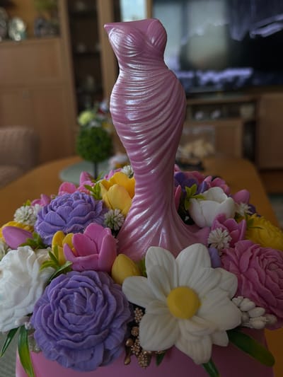 A close-up, eye-level, indoor shot showcases a decorative pink cake shaped like a flowing dress, adorned with an assortment of intricately crafted soap flowers in vibrant shades of purple, pink, yellow, and white. The soap flowers are arranged around the base of the dress-shaped cake. The background is blurred, suggesting an indoor setting with furniture and possibly a television. The lighting is soft, highlighting the pearlescent sheen of the dress cake and the delicate details of the soap flowers. There is no indication of the city of Lyubimets, Bulgaria, in this image.