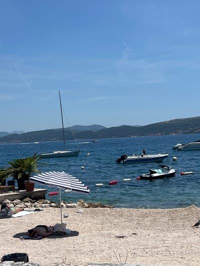 The image depicts a sunny day scene at a beach in Baošići, Montenegro. In the foreground, a sandy beach is visible with two individuals sunbathing. One person is lying on their back under a blue and white striped umbrella, while another person is lying on their stomach on a towel further back.
The water in the background is a deep blue, with several boats visible. A sailboat with a tall mast is anchored offshore, and several smaller motorboats and a jet ski are also present. A line of buoys separates the swimming area from the boats. In the distance, rolling green hills covered with vegetation and scattered buildings form the coastline. The sky is clear blue with a few faint contrails from airplanes. The overall atmosphere is one of leisure and relaxation on a summer day.