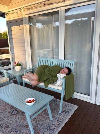 A woman is sleeping on a light blue bench on a wooden deck. She is covered with a green blanket and has a patterned pillow under her head. The bench has cushions. In front of the bench is a light blue coffee table with a white bowl of red fruit. A patterned rug is on the deck under the table and bench. A small side table with a potted plant is to the left of the bench. In the background are large glass doors and siding of a house. String lights are visible above the doors. The location is described as Starokorsunskaya, Russia.