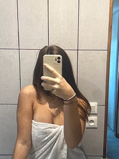 A person, identified as female based on visual cues, is captured in a mirror selfie. She is positioned in what appears to be a bathroom, indicated by the tiled wall and electrical outlets visible behind her. The individual is wrapped in a light-colored towel, covering her lower body. Her upper body is partially exposed, and she is wearing a delicate necklace. Her right arm is raised, holding a smartphone to take the photograph, with her fingers obscuring her face. A bracelet is visible on her right wrist. The background consists of textured, light-colored tiles arranged in a grid pattern, a wooden door frame, and electrical outlets. The overall lighting suggests an indoor environment. The scene does not contain any identifiable elements or landmarks that would suggest a specific location within Pruszcz Gdański, Poland.