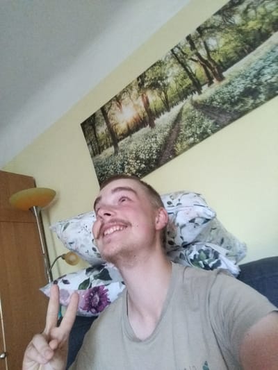 This is a close-up, low-angle, medium shot of a young person, likely male, making a peace sign with their left hand. They have short, light brown hair, a light mustache, and are smiling with their mouth open, revealing teeth. They are wearing a light-colored t-shirt. The person is leaning back against several pillows, one of which has a floral pattern with a large purple flower. In the background, a large landscape artwork of a sunlit forest path with white flowers is hanging on a pale yellow wall. To the left of the person, a floor lamp with an orange shade is visible, as well as a portion of a wooden closet door with a silver knob. The scene appears to be indoors, possibly a bedroom or living room. The lighting suggests it could be daytime. The person's expression and gesture convey a sense of happiness and friendliness. There is no visible text in the image. The location context is Cēsis, Latvia.