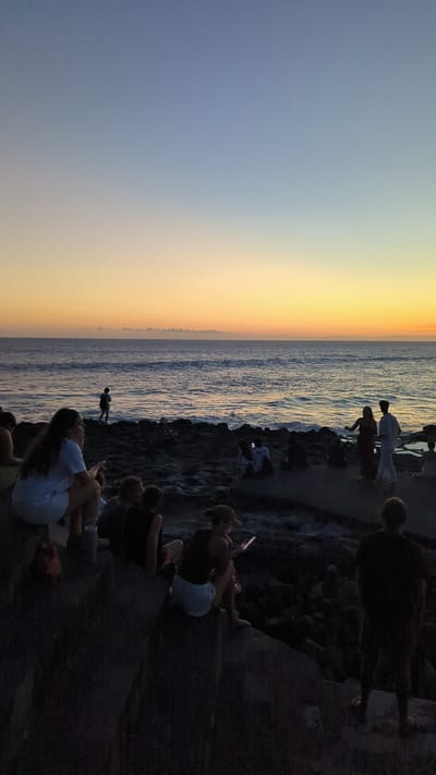 The image shows a group of people gathered at a beach in Pererenan, Indonesia, possibly during sunset. To the left, the sky transitions from blue to orange, with the sea below. On the right, people are sitting and standing on what appears to be a rocky outcrop or shoreline. Some individuals are observing the sunset, while others are looking at their phones. There is a person wading in the water. The overall scene suggests a relaxed, social gathering during a scenic sunset.