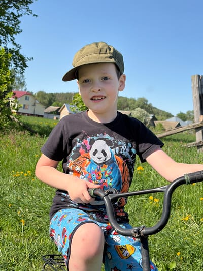 A young boy is outside on a sunny day in what appears to be a grassy field. He is wearing a cap, a black t-shirt with a graphic design featuring a panda and a tiger, and blue shorts with baseball-themed cartoon designs. He is holding onto the handlebars of what appears to be a tricycle or similar wheeled vehicle. In the background, there are houses, trees, and a blue sky, indicative of a residential or rural setting. The scene suggests a leisurely, playful activity in a natural outdoor environment. The location could be Кемуль, Russia, given the prompt's specification.