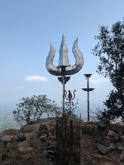 The image displays a rocky hilltop location in Kadaladi, India, under a clear blue sky. The central subject is a large, metallic trident (Trishula) constructed from silver-colored metal plates, positioned upright. To its right, a tall, dark metal pole with a flat, cross-shaped top stands beside a dark-leaved tree partially visible on the right edge. In the midground, a smaller, rusty metal structure resembling a trident, adorned with multiple small bells, is surrounded by sparse green vegetation within a protective wire frame. The ground is dry and uneven, covered with rocks and reddish-brown soil, featuring several cairns (stacks of rocks). On the left, a tree with yellow flowers grows on a rocky prominence. The distant background reveals a hazy panoramic view of a valley with cultivated fields and scattered buildings, indicating a significant elevation. No people are visible.