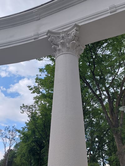 The image is a close-up, eye-level shot of a white, textured, fluted column with an ornate capital. Above the column, a white architectural element curves into the frame, featuring decorative trim. The background is a mix of lush green trees and a blue sky with scattered white clouds. The location is identified as Alikonovka, Russia, though the specific setting within the city is not discernible from the image alone. There are no people or animals visible in the frame. The lighting suggests it is daytime, possibly overcast due to the diffused light.