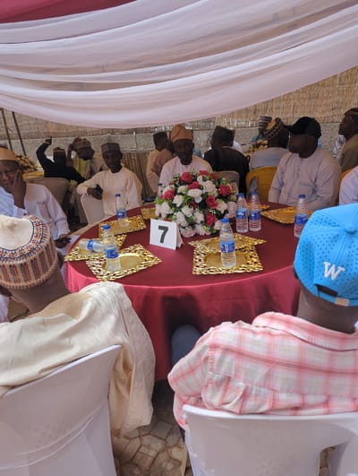 This image depicts a gathering of approximately 10-15 men under a temporary canopy structure, taking place in Kabala, Nigeria. The scene centers around a round table covered with a dark red tablecloth, equipped with a floral centerpiece of red and white roses.
**People:**
In the immediate foreground, two men are seated with their backs partially facing the camera. The man on the left wears a light-colored traditional garment with a patterned cap featuring brown, orange, and white hues. His right arm rests on a white chair cover. The man on the right wears a light blue baseball cap with a white 'W' logo on the back and a pink and white plaid shirt; his left shoulder is visible, also resting on a white chair cover.
Around the round table, multiple men are seated. A man directly opposite the viewer wears a light-colored traditional outfit and matching cap, looking forward. To his left (viewer's right), a man in a light-colored striped shirt and a black baseball cap is seated, facing left. To his right (viewer's left), another man in a dark traditional outfit and cap is partially obscured, with his hand near his face. Further back and to the left, a man is visible with his right arm raised and index finger pointing upwards. The majority of individuals are dressed in traditional Nigerian attire, including long robes and various caps, while some wear Western-style clothing. Several men have mobile phones in their hands or placed nearby.
**Objects and Setting:**
The red-covered table features multiple ornate, gold-colored rectangular trays, each holding a clear plastic bottle of "CWAY" brand water. A white tent card displaying the number "7" in black is positioned on the table to the right of the floral centerpiece. The chairs are covered with white fabric. The gathering is situated under a white fabric canopy, draped in gentle folds across the upper portion of the frame, providing shade. In the background, behind the seated individuals, a light-colored wall or partition made of woven, natural material (possibly bamboo or reeds) is visible. The ground beneath the chairs is paved with irregular, light-toned stones or tiles. The scene is illuminated by natural daylight.