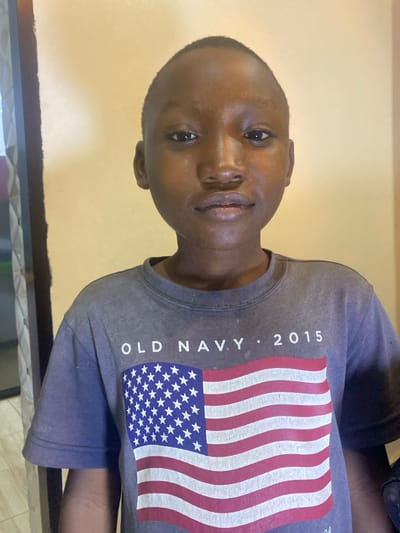 A young Black boy with close-cropped hair is depicted from the chest up. He has brown skin, and his face is turned slightly toward the viewer. He's wearing a gray t-shirt with a print of the American flag on the front. The words "OLD NAVY" and "2015" are also visible on the shirt. The background appears to be a light yellow or beige wall. The image is rotated counterclockwise, placing the wall at the left and the boy at an angle.
