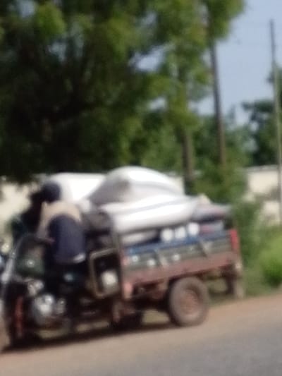 The image shows a motorcycle with an attached wagon moving along a paved road. The motorcycle is carrying a load covered by a white tarp. A person is riding the motorcycle. The background includes trees, poles, and a building. The location is presumed to be in the city of Dungu, Ghana.