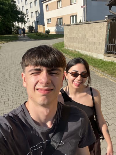 This is a vertical, medium shot selfie taken outdoors on a sunny day. The image features two young people. In the foreground, a young man with short, dark brown hair smiles and looks towards the camera. He is wearing a dark t-shirt with a white graphic design on the sleeve. To his right and slightly behind him, a young woman with dark hair and sunglasses sticks out her tongue. She is wearing a dark tank top.

The background shows a paved walkway and a grassy area. Beyond that, there is a modern apartment building with cream and beige facades and numerous windows. In the distance, two people can be seen walking. The lighting suggests it is daytime, likely afternoon, given the strong shadows. The overall mood appears lighthearted and casual. There is no visible text in the image. The location context provided is Dobřany, Czechia.