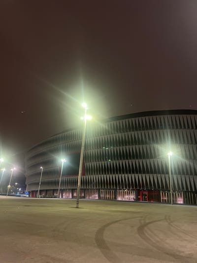 The image shows the San Mamés Stadium, located in the city of Bilbao, Spain. It is nighttime, and the stadium's exterior is illuminated by several streetlights, casting a bright glow. The stadium has a distinctive modern design with a curved facade covered in triangular panels. The surrounding area appears to be a large, open plaza or parking lot, with tire tracks visible on the ground. The sky is dark and overcast, with some atmospheric haze. There are no people visible in the image.