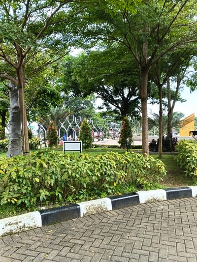 This image displays a section of a public park or green space located in the city of Bien Hoa, Vietnam. The foreground features a herringbone-patterned paved pathway or surface, bordered by a concrete curb painted with alternating black and white sections. Immediately behind the curb, a dense bed of variegated shrubs with green and yellow foliage is present.
The midground is characterized by a manicured green lawn, dotted with numerous trees of varying sizes, all featuring dense green canopies. A prominent white rectangular sign with a black border, supported by two green poles, stands centrally on the grass. Several conical evergreen shrubs are also interspersed within the grassy area.
In the background, a sequence of white, ornate, wave-like architectural structures extends horizontally into the distance, possibly forming a decorative arcade or pergola. Beneath and around these structures, several indistinct human figures are visible, appearing to be stationary or in casual movement within the park environment. On the far right, a building with a yellow or golden-colored roofline is partially visible beyond the trees. The overall environment is lush with vegetation under a bright, possibly overcast, sky. No specific actions or interactions among the distant figures are discernible due to resolution.