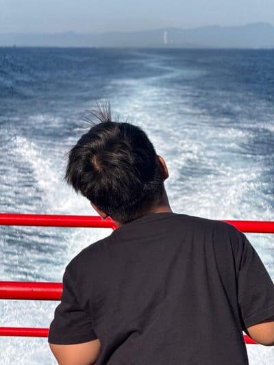 A young person wearing a black t-shirt is leaning their head back, looking out at the ocean from a boat. Red railings are visible in the foreground. The water behind them shows a churned-up wake. In the distance, the hazy silhouette of mountains and a tall, thin structure are visible on the horizon. The location is identified as Dong Son, Vietnam.