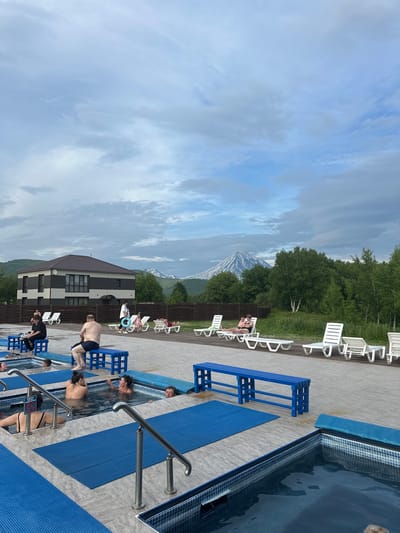 The image depicts an outdoor hot spring facility with several people relaxing in pools. The setting is located in Yuzhnyye Koryaki, Russia, with a prominent volcano visible in the background under a cloudy sky. In the foreground, there are multiple rectangular hot tubs filled with water, some with people bathing in them. Blue mats and blue benches are scattered around the tiled ground. Further back, there is a building with a dark roof and light-colored walls, and a wooden fence. White lounge chairs are arranged on a wooden deck. The overall atmosphere is one of leisure and relaxation.
