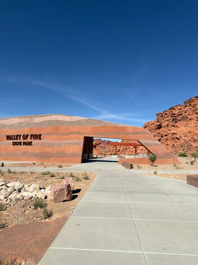 The image depicts the entrance to Valley of Fire State Park in Moapa Valley, Nevada, United States. The park's name is prominently displayed on the building, which has a striped pattern of alternating colors. There's a blue sky visible in the background. The building extends into the landscape on one side. There is a red rock outcrop on the other side, and a concrete sidewalk adjacent to the building. Small patches of desert vegetation are visible.