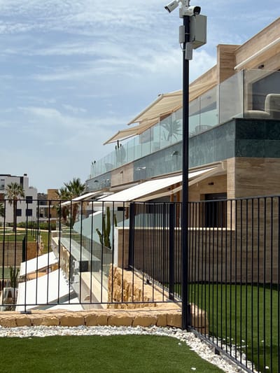The image depicts a modern building with multiple balconies and awnings, situated in Finestrat, Spain. A black metal fence runs across the foreground, partially obscuring a meticulously maintained green lawn interspersed with white pebbles. Behind the fence, the building features a façade of light-colored travertine stone and dark green marble accents. Glass railings line the balconies, and several retractable awnings are deployed. A lamppost with two security cameras is visible on the left side of the image, with the building's entrance and windows visible in the background, including palm trees and other vegetation. The sky is partly cloudy, indicating daytime.