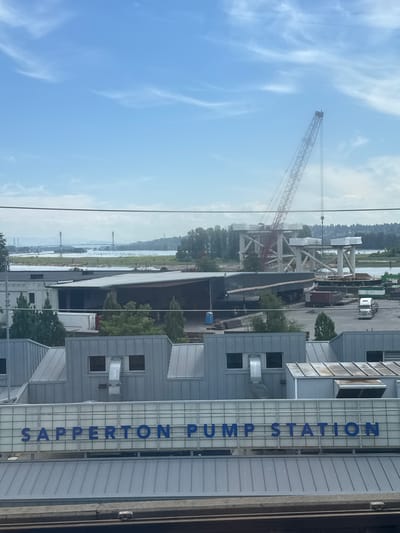 The image shows a view from a train, looking out at a waterfront industrial area. In the foreground, there is a building with "SAPPERTON PUMP STATION" written in large blue letters across its facade. Above the building, the sky is blue with some wispy clouds. A large construction crane is visible in the middle ground, along with the framework of a bridge under construction. Beyond that, a wide river is visible, with a distant shoreline and mountains. The overall scene suggests a working port or industrial zone in an urban or semi-urban setting. The location is identified as New Westminster, Canada.