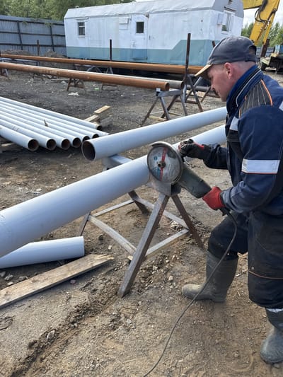 This image captures a man working outdoors, likely in an industrial or construction setting in Parma, Russia. The subject, a man wearing a blue and black work uniform, a gray cap, and red gloves, is actively using a grinder to cut a large, light gray metal pipe. Sparks are flying from the point of contact, indicating the intensity of the work. He is wearing dark work pants and black rubber boots.

The scene is set on rough, dirt ground scattered with small rocks and debris. Several large metal pipes, both rusted brown and freshly painted light gray, are visible in the background, suggesting a construction or fabrication project. Some of these pipes are supported by metal stands. In the far background, a white and light blue prefabricated building or trailer is partially visible, with a yellow construction vehicle behind it. The overall impression is one of active, manual labor in a somewhat rugged environment.

The lighting suggests it is daytime, with sunlight casting shadows. The weather appears to be fair. There is no discernible text in the image. The man's focused expression and the act of grinding suggest a task requiring concentration and precision.