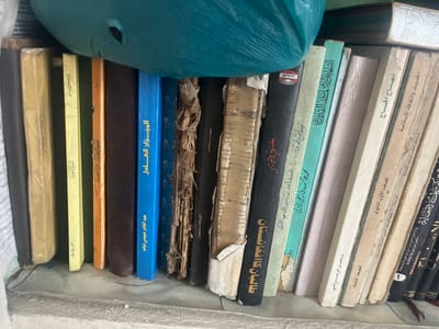 The image shows a close-up, eye-level view of a bookshelf filled with old, bound books, mostly in Arabic. The books are stacked tightly, with some leaning against each other. The spines of the books display titles in Arabic script. Some of the book covers are faded and worn, indicating their age. The shelf appears to be made of concrete or a similar rough material. In the upper part of the image, a large, crumpled teal-colored plastic bag is visible, partially obscuring the tops of the books. The lighting suggests an indoor environment, possibly a storage area or a dimly lit room. The overall scene conveys a sense of disarray and neglect. There are no people or animals present in the image. The location is described as Tashlak, Kyrgyzstan.