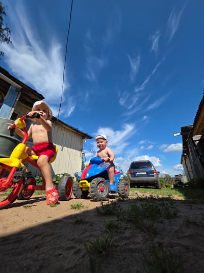 Two young children are on toy vehicles in a rural area, possibly in Буренка, Russia, on a sunny day with wispy clouds. The child on the left is on a red and yellow tricycle, wearing red shorts and a white hat. The child on the right is on a blue and yellow ATV-style toy, also wearing a white hat. In the background, a blue SUV is parked near a wooden fence and a rustic building. The ground appears to be dirt and sand with some sparse green vegetation. The perspective is low-angle, emphasizing the sky and the children's perspective.