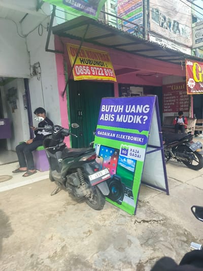 The image shows a man wearing a black hooded jacket and a face mask sitting on a bench looking at his phone, with a dark green motorcycle parked behind him, in front of a pink storefront. The store is located at -6.543, 107.441 in Purwakarta, Indonesia. A large green and yellow sign is situated next to the motorcycle, advertising an electronics pawn shop with the text "Butuh Uang Abis Mudik?" which translates to "Need money after Eid?" and the phrase "Gadaiakan Elektronik!", meaning "Pawn Your Electronics!". Another sign above it states "Over Kontrak" and displays a phone number. There is a sign to the right of the man on the bench offering salon services with a price list for services. The motorcycle's license plate is visible. The sidewalk is cracked and grey. The sky above the storefront is cloudy.