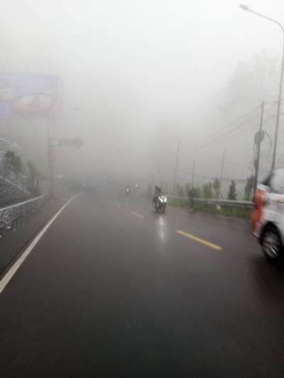 A foggy road scene shows limited visibility. A billboard is visible on the left side of the road. Several motorbikes are seen riding on the road, with a car visible on the right. The road is wet, suggesting recent or current rain. Yellow lane markings are present. Utility poles and a roadside barrier are visible on the right. The heavy fog obscures distant features.