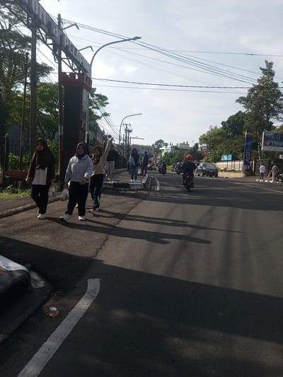 The image depicts a street scene in an urban area, possibly in Asia, based on the appearance of the people and environment. A group of four women are walking along the sidewalk on the left side of the road. They are dressed in casual clothing, some wearing headscarves. One of the women has her arm raised.
On the right side of the road, there are motorcycles and cars driving. There are other people walking and standing along the sidewalk.
Overhead, there are power lines. Trees and buildings are visible in the background. A sign with the word "PELOPOR" is prominent on the left side of the image. The ground is paved and appears to be dry. The light suggests it is daytime.