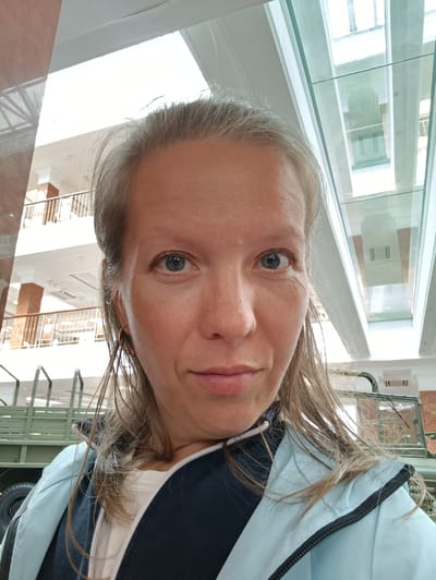 A woman with blonde hair and blue eyes takes a selfie in front of a museum exhibit. She is wearing a light blue and dark blue jacket over a white shirt. Her hair is pulled back from her face. In the background, there is a display of military vehicles, including a green truck, and architectural elements of the museum, such as railings and glass enclosures. The museum appears to be the Military Technology Museum "Battle Glory of the Urals" in Verkhnyaya Pyshma, Russia, as evidenced by the exhibits. The lighting suggests an indoor setting with natural light coming from above.