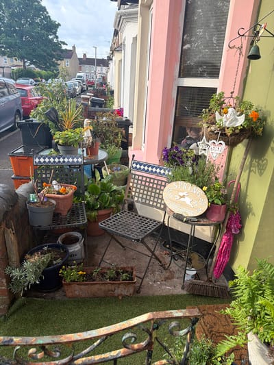 This image shows a street scene in Swindon, United Kingdom. The focus is on the front garden or walkway of a terraced house. The houses are close together, and the street appears narrow with cars parked tightly along the roadside. The house features a pink facade on a portion of the building.
The area is filled with a collection of potted plants, garden ornaments, and small pieces of furniture, including a small table and chairs. There are various types of plants, including flowers and greenery, in pots of different sizes and materials. Garden decorations, such as a bunny-themed welcome sign, angel statue, and other decorative objects, are interspersed among the plants. There is a visible broom, rake and pink umbrella as well.
The foreground shows a mix of hardscaping elements, like brick and concrete. Artificial turf is visible along a section of the boundary. Weathering and wear are evident on the buildings and objects, suggesting a lived-in urban environment.
No people are visible in the image.