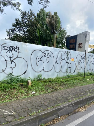 The image shows a white concrete wall with graffiti on it. The wall is located along the side of a paved road. The road is lined with lush green vegetation. The coordinates indicate a location in Lembang, Indonesia, near a building with a sign that reads "Grand Hotel." The wall is covered with black spray paint in various abstract shapes and letters, such as "678" and "O." The wall appears to be a construction barrier, and the "Grand Hotel" sign suggests that the construction is related to a hotel project. There is a large sign on a metal pole behind the barrier. The sign on the metal pole says "Jl. Terbata A". The scene is captured from a moving vehicle, providing a slightly blurred perspective.