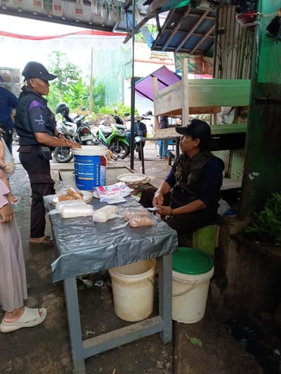 This image captures an outdoor street vendor scene, likely in an informal market in Ciamis, Indonesia. In the foreground, a simple wooden table with a black plastic cover displays various goods wrapped in clear plastic, possibly food items like snacks or raw ingredients. Two white plastic buckets, one with a green lid, sit beneath the table.

Two main individuals are featured. On the left, a person stands, wearing a black cap, a dark vest over a dark short-sleeved shirt, and dark cargo pants with sandals. They are positioned near a large blue bucket with a white lid, which bears the text "3 IN 1" and some Chinese characters. Their hands are resting on or near the bucket. Partially visible on the far left are the legs and lower torso of another person. On the right, a second person is seated on a low wooden stool, also donning a black cap and a dark vest over a long-sleeved shirt. This individual, presumably the vendor, is looking away from the camera.

The background reveals a bustling, semi-urban environment. Several motorcycles, including scooters, are parked. Simple market stalls constructed from wood and corrugated metal, some with tarps, line the area. White sacks with the letters "SR" repeatedly printed on them hang overhead, possibly serving as a canopy or part of a display. Lush green vegetation is visible further in the background, contrasting with the man-made structures. The ground is a mix of concrete and packed earth.

The scene depicts a daily activity in a local market, with goods laid out for sale. The lighting suggests it is daytime, possibly overcast or shaded, as there are no harsh shadows. The overall atmosphere appears casual and routine, with no strong emotions discernible from the subjects.